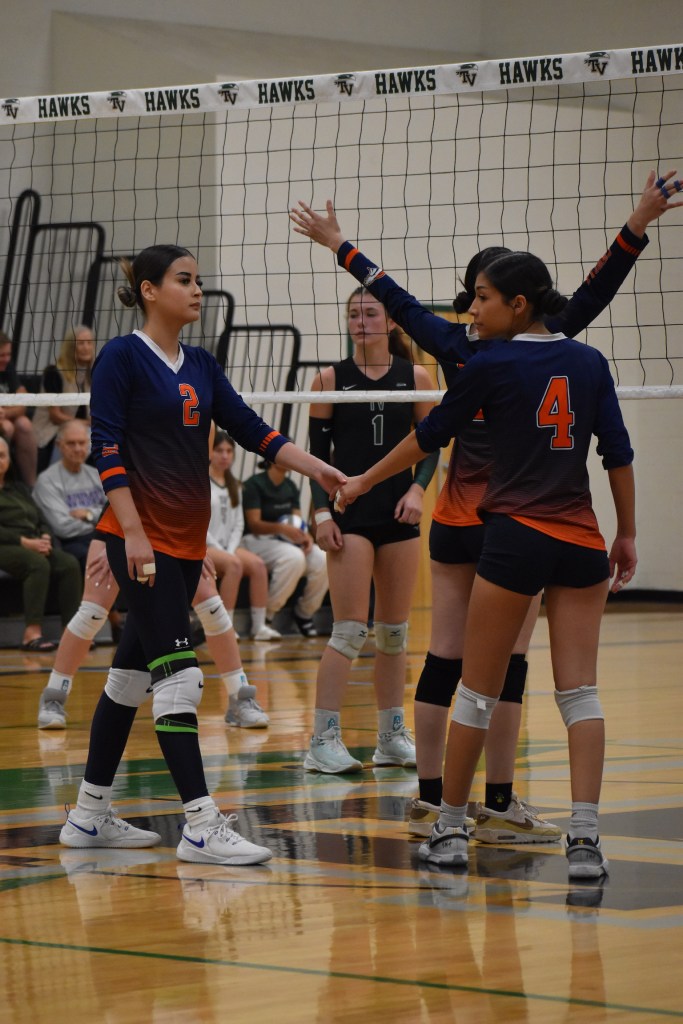 Two Varsity players holding hands on court and Middle blocker hands up on net getting ready for their player to serve 