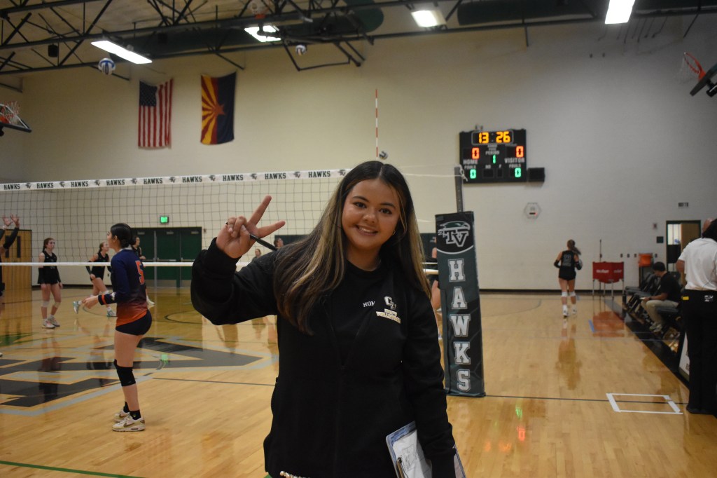 Young Lady (The Volleyball Coach) holding up a piece sign and smiling, wearing all black with clipboard in arm, on court 