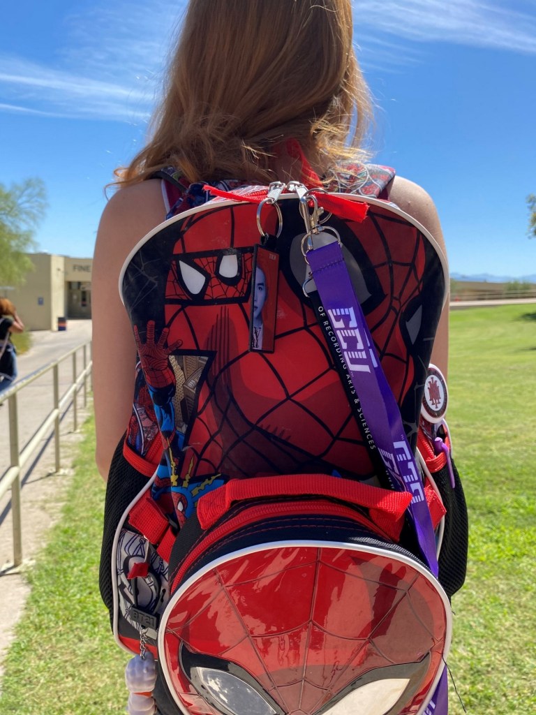 Girl outside in front of grass with back facing the camera to show Spiderman backpack. 