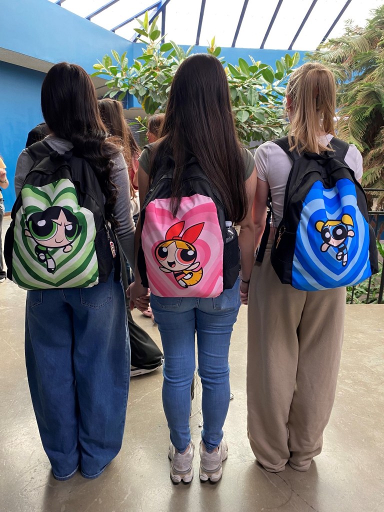 Three girls posing with their backs facing the camera to show their matching backpacks. From left to right the bags have the characters, Buttercup, Blossom, and Bubbles. 