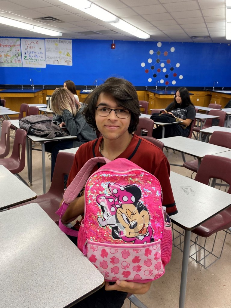 Boy front-facing while sitting at a desk in a classroom holding up Minnie Mouse backpack  